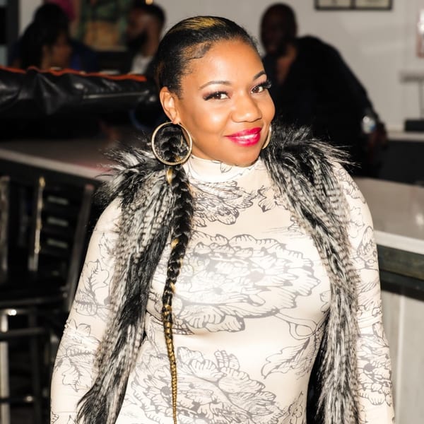 Woman in a patterned dress and faux fur vest, with a long braid and hoop earrings, smiling at Empire Lounge.