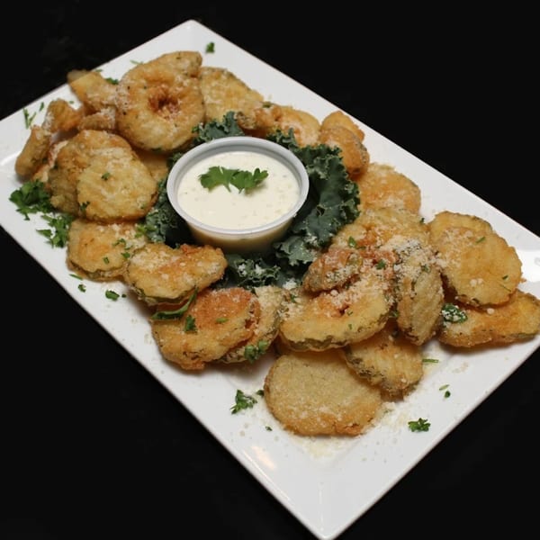 A white rectangular plate filled with golden-brown fried pickles, garnished with parsley, cheese, and a side of ranch.