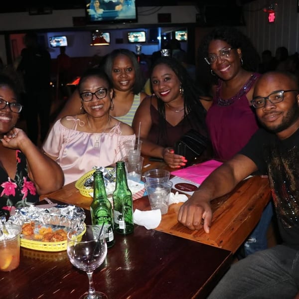 Six smiling patrons, four women and two men, seated around a table with drinks and food at Empire Lounge.