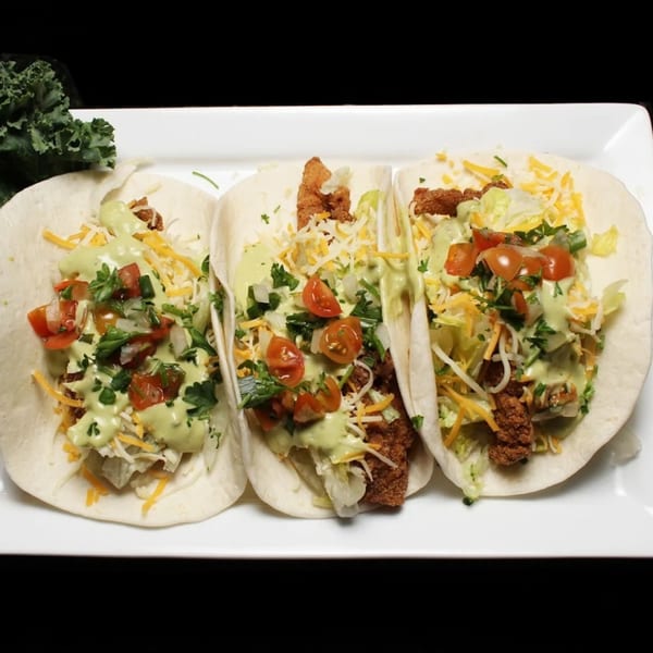 Three Empire Lounge tacos with fried chicken, shredded lettuce, cheese, pico de gallo, and green sauce.