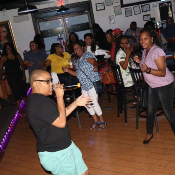 A person sings karaoke into a microphone while a group of people dance and cheer in a bar.