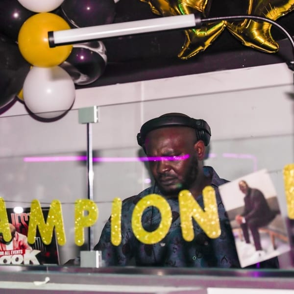 DJ with headphones on, behind a glass barrier with "CHAMPION" in glitter letters.