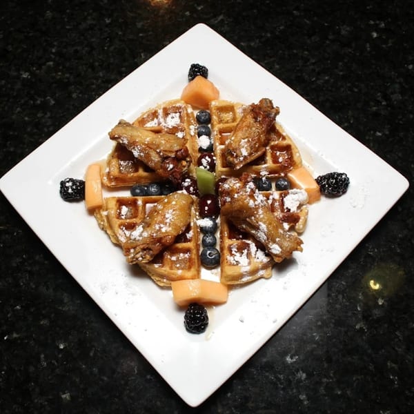 Chicken and waffles with powdered sugar, blueberries, blackberries, and cantaloupe on a white plate.
