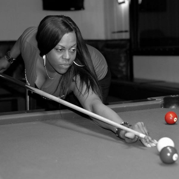 A woman in a bar lines up a shot on a pool table, with a red ball highlighted.