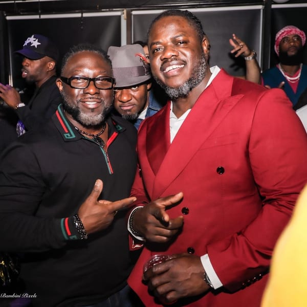 Two men smiling at the camera, one in a red suit, the other in a black polo shirt.