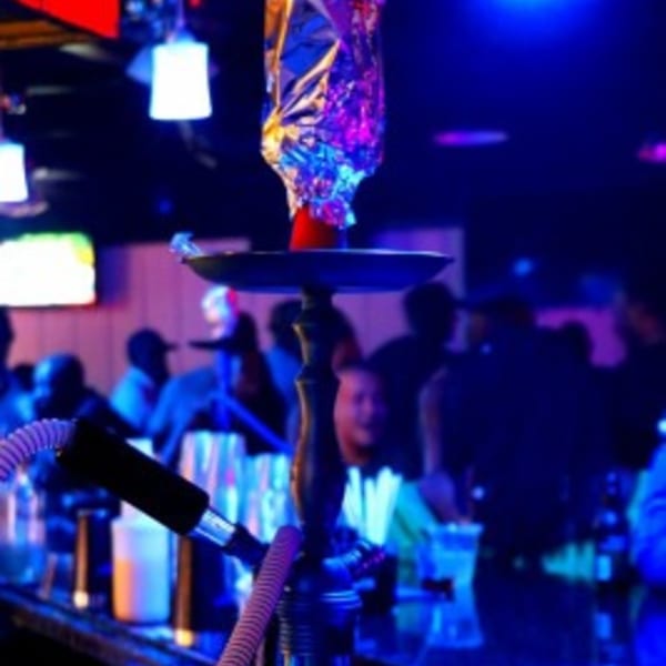 A hookah with foil-covered coals sits on a bar in a dark lounge with blue lighting and blurred patrons.