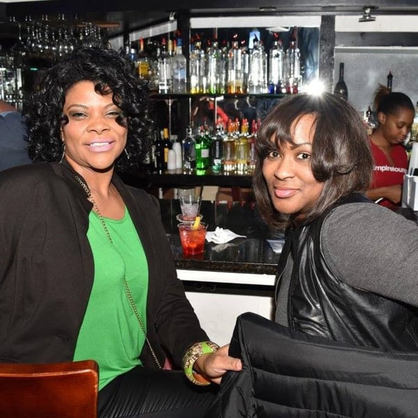 Two women smiling at the camera, seated at a bar with drinks and bottles in the background.