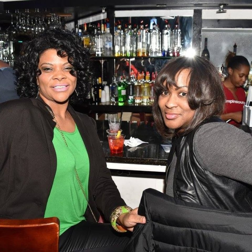 Two women smiling at the camera, seated at a bar with drinks and bottles in the background.